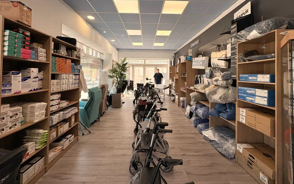 Allée centrale du magasin de Miramas : fauteuil releveur turquoise, rollators alignés et rayon confort, avec la lumière de la vitrine au fond